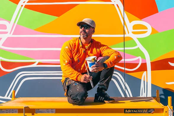 A male artist, wearing an orange paint-splattered shirt and cap, crouches on a yellow platform in front of a colorful mural with geometric shapes and white outlines of vehicles. He holds a paint can and brush.