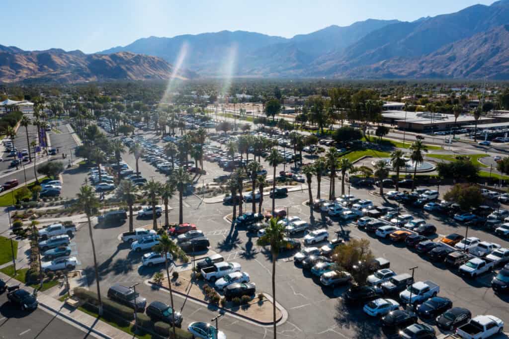 Aerial view of multiple parking lots full of cars and palm trees, with a large mountain range in the background under a clear sky.