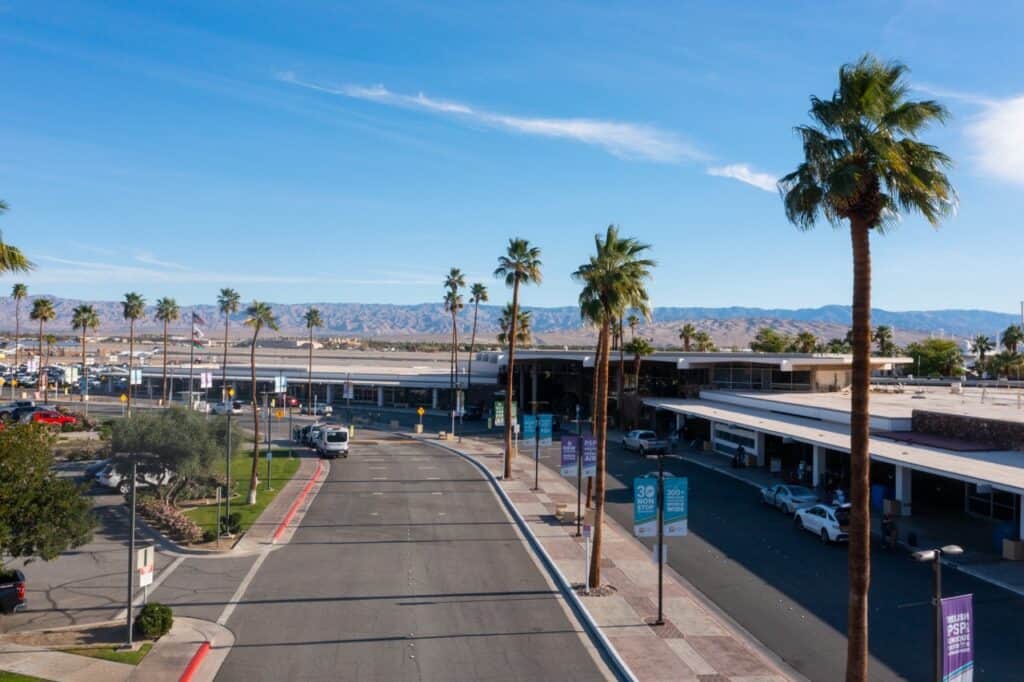 Elevated view of Palm Springs International Airport with palm trees, roads, terminal buildings, and distant mountains under a blue sky.