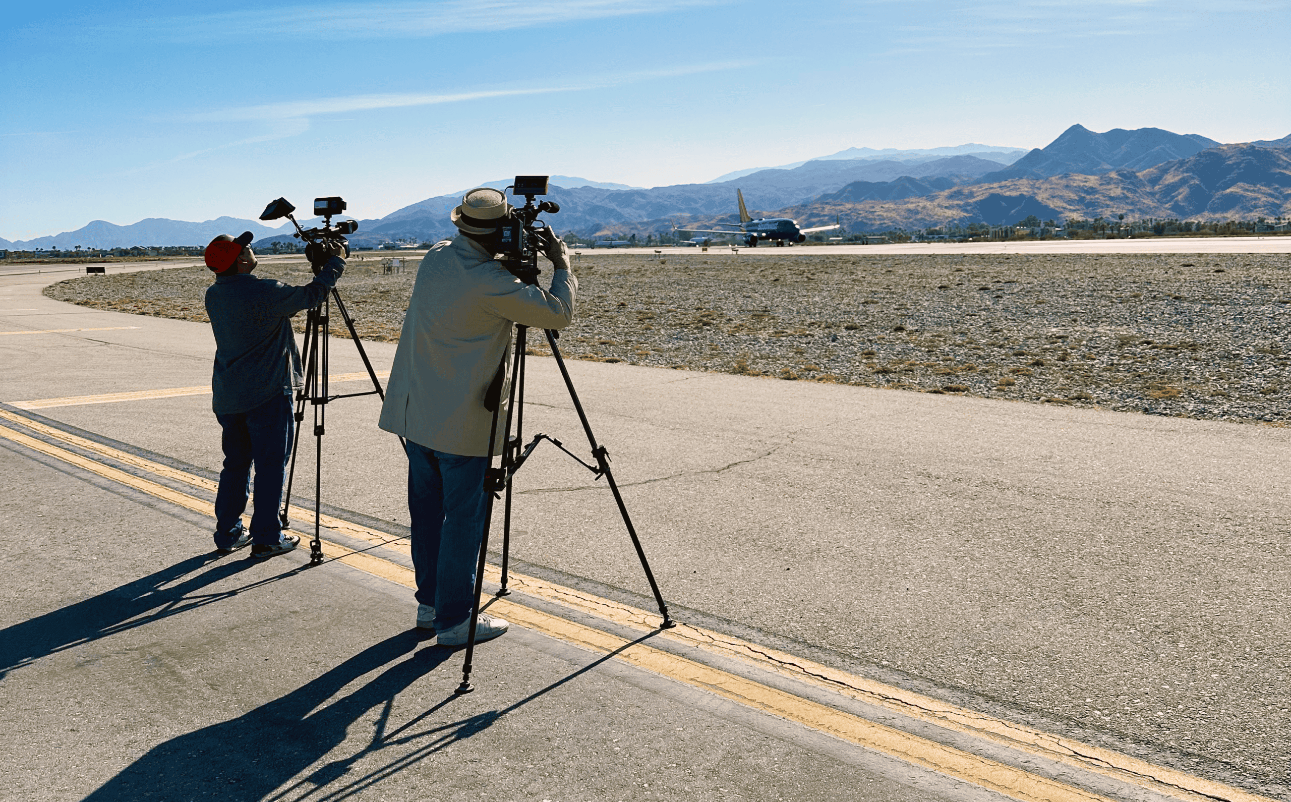 Two men with video cameras on tripods film a blue Southwest Airlines plane on an airport runway, with mountains in the background.
