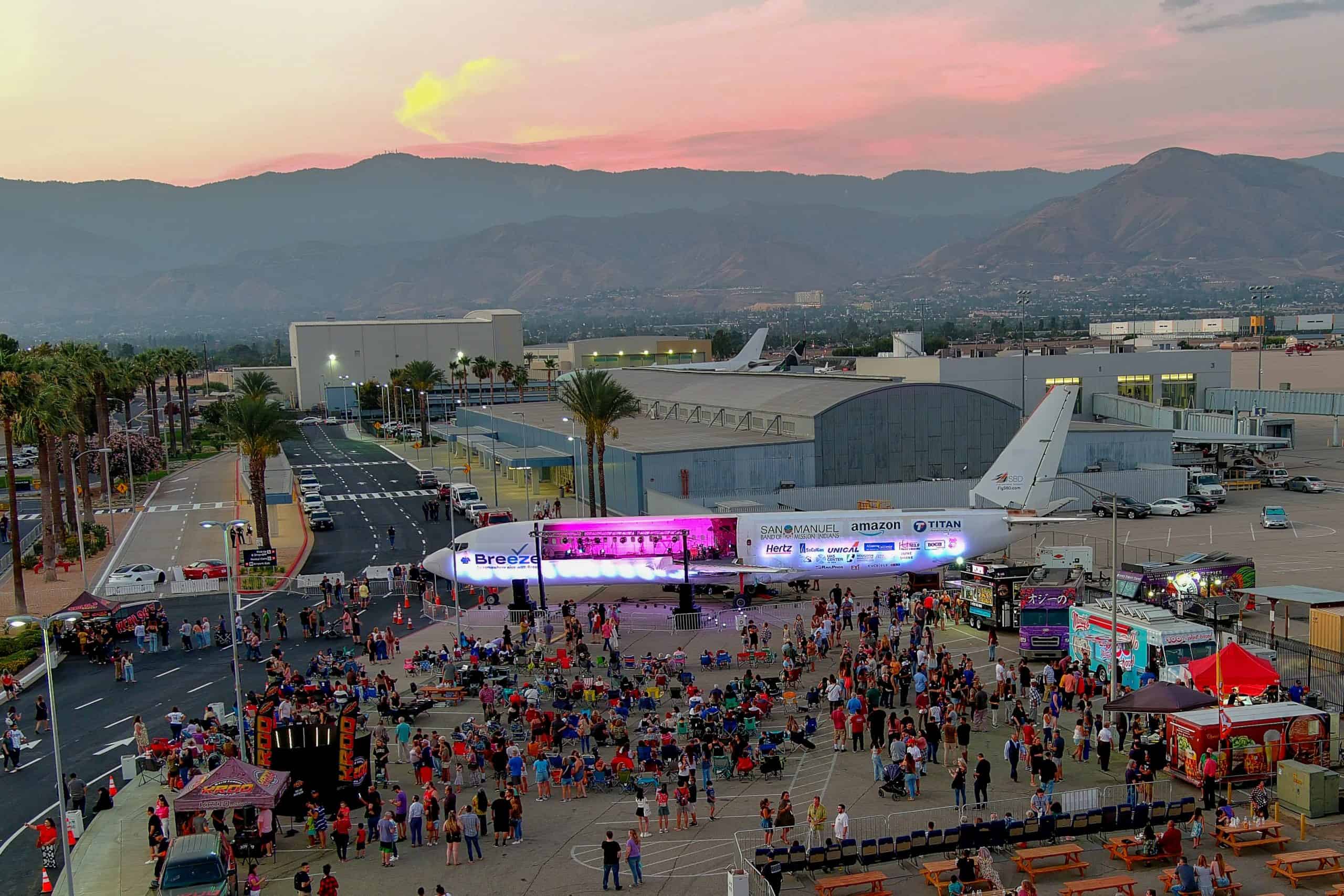 Aerial view of the Cruising at Altitude festival grounds on the SBD tarmac at sunset, with food trucks, a large screen and crowds gathered around the Plane Stage.