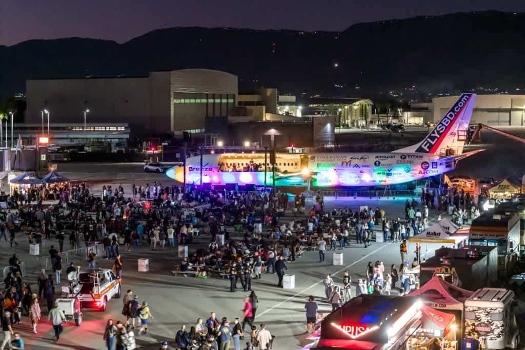 Aerial view of the festival grounds in the evening, with a parked jet, food trucks and crowds on the tarmac.