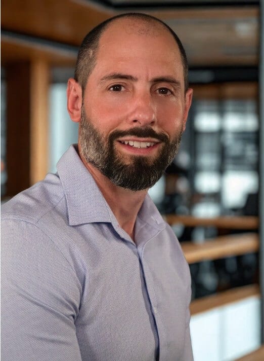 A man with a trimmed beard and short hair, wearing a light purple button-up shirt, stands indoors in a modern office with wooden and glass details, smiling at the camera.