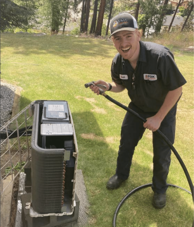 A technician wearing a uniform and hat is smiling while holding a hose and cleaning an outdoor air conditioning unit on a lawn.