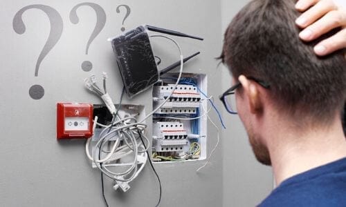 A man looks confusedly at a messy network of wires, routers, and switches exposed in a wall panel, wondering if calling a local electrician was the right choice—question marks float in the background.