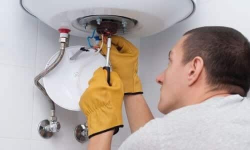 A person wearing yellow gloves uses a screwdriver to repair plumbing components under a water heater, possibly diagnosing issues related to furnace repair or a broken heater.