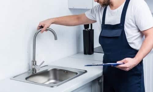 A person in work overalls checks a kitchen faucet while holding a clipboard next to a stainless steel sink, illustrating the plumbing inspection purpose.