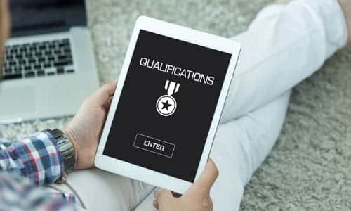 Person holding a tablet displaying the word "QUALIFICATIONS" with a medal icon and an "ENTER" button, seated on the floor near a laptop—highlighting the importance of checking plumber qualifications before hiring a plumber.