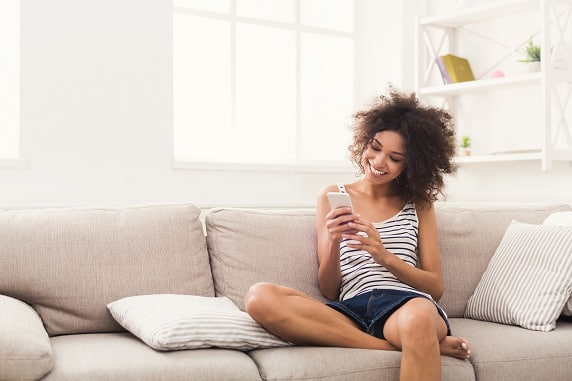 Woman sitting on a couch, smiling while looking at her smartphone, with one leg crossed and a striped pillow beside her—perhaps enjoying an exclusive Newsletter Sign-Up offer.