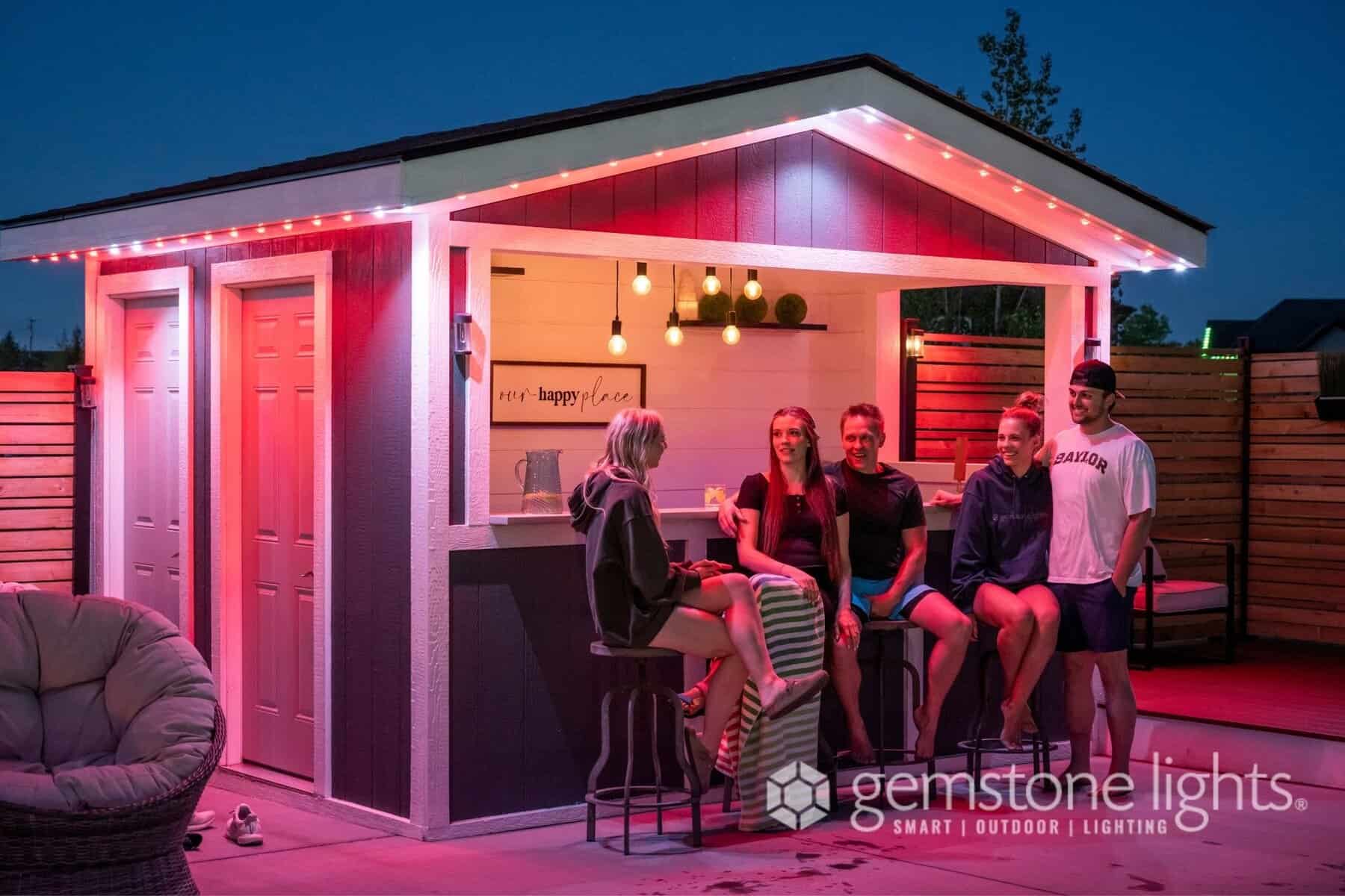 Five people sit and stand around a modern outdoor patio bar at dusk, illuminated by red LED lights along the roof, with an "about us" gemstone lights logo in the corner.