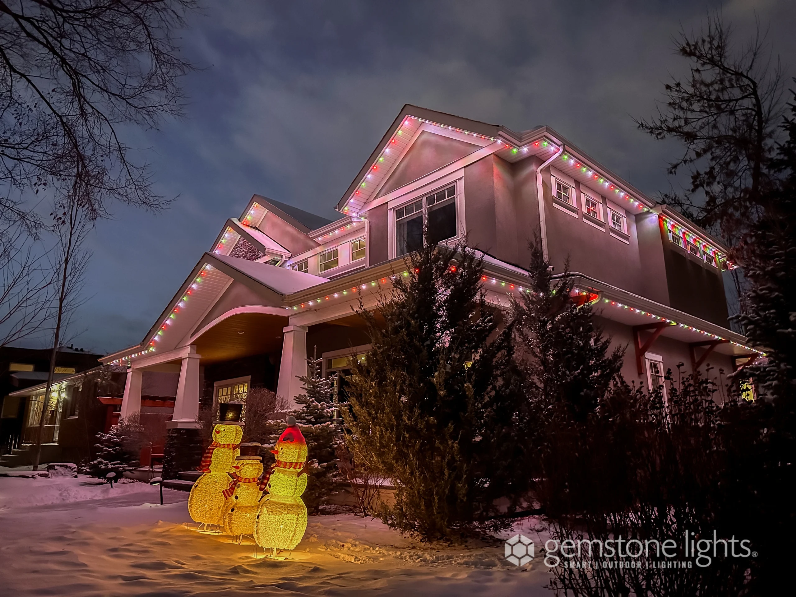 A two-story house decorated with colorful holiday lights along the roofline, with two illuminated snowman figures on the snowy lawn at dusk, perfectly captures the warmth and spirit you'll find in our "about us" story.