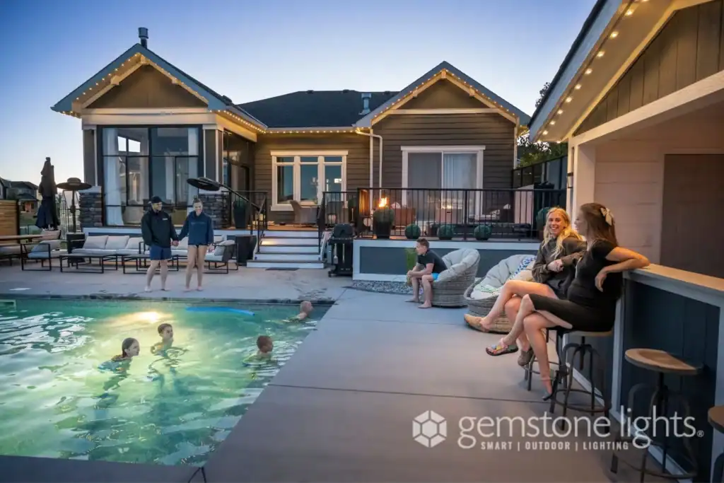 People gather around a backyard pool at dusk; some are swimming while others sit and talk near the pool and at the bar, reflecting the relaxed, welcoming spirit that defines about us. A house with outdoor lights glows in the background.
