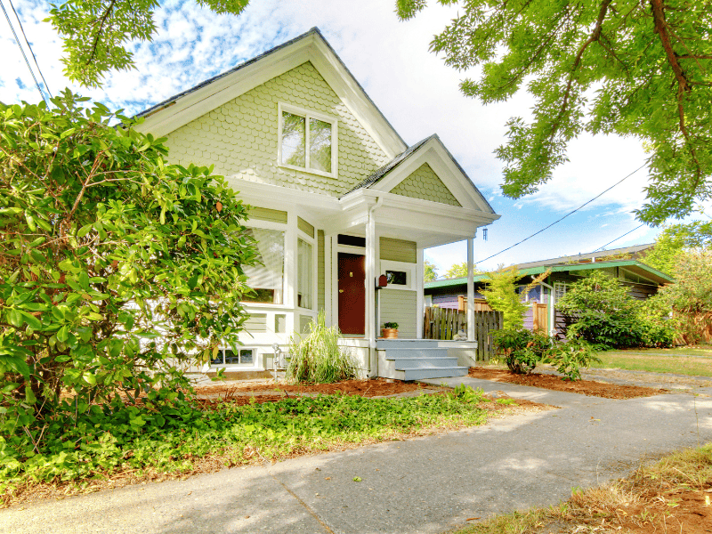 A light green house with a gabled roof and a front porch, surrounded by greenery and trees, sits on a paved walkway under a partly cloudy sky. It's prepped for the summer season with efficient AC keeping it cool amidst the warmth.