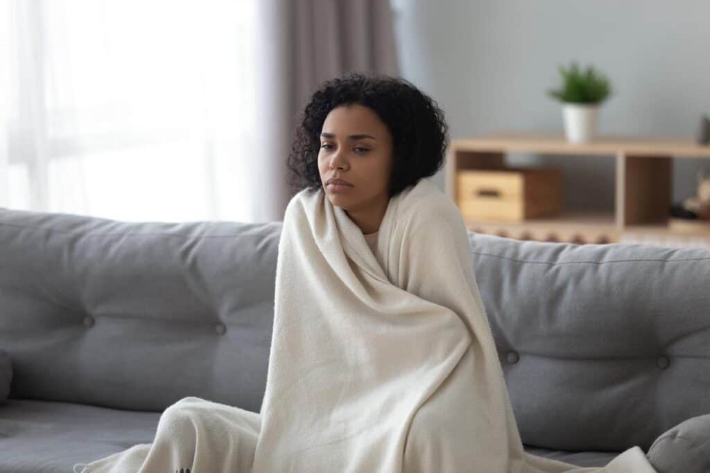 A person sits on a gray couch wrapped in a blanket, looking thoughtful as the heater blows warm air. A shelf with a plant and boxes is in the background.