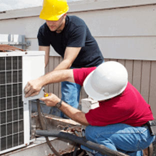Two workers in hard hats are installing or repairing an outdoor air conditioning unit. One wears a yellow hat, black shirt, and the other a white hat, red shirt.