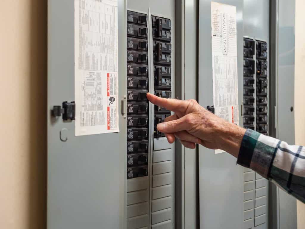 A hand pointing at switches on an electrical breaker panel with labeled diagrams attached to the door highlights essential benefits, emphasizing the importance of regular service inspections.