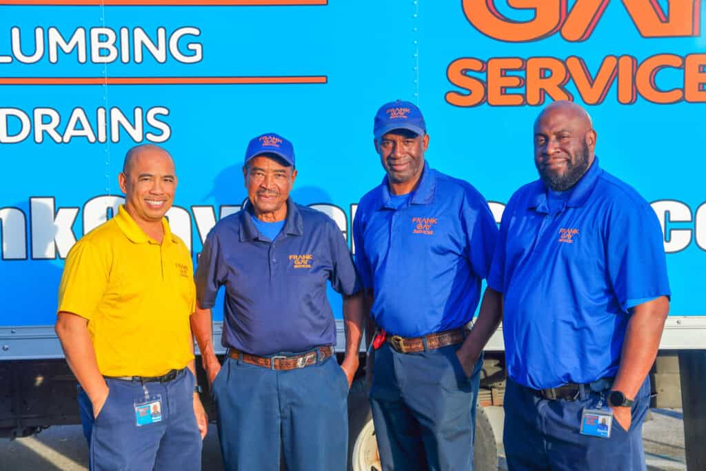 Four men in uniforms stand proudly in front of a blue plumbing and drain service truck, showcasing their careers. Three sport blue shirts and caps, while one stands out in a yellow shirt.