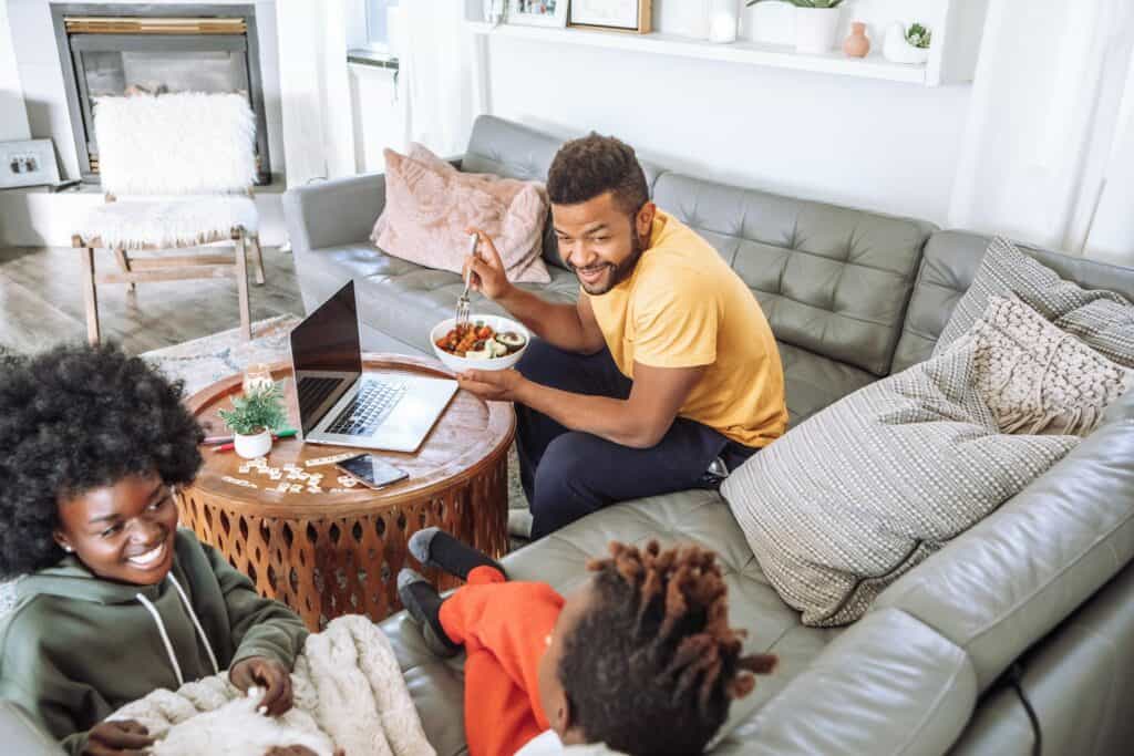 A man sits on a couch eating from a bowl and smiling at two children. An open laptop, a puzzle, and smart filters for air filtration are on the coffee table in a bright living room.