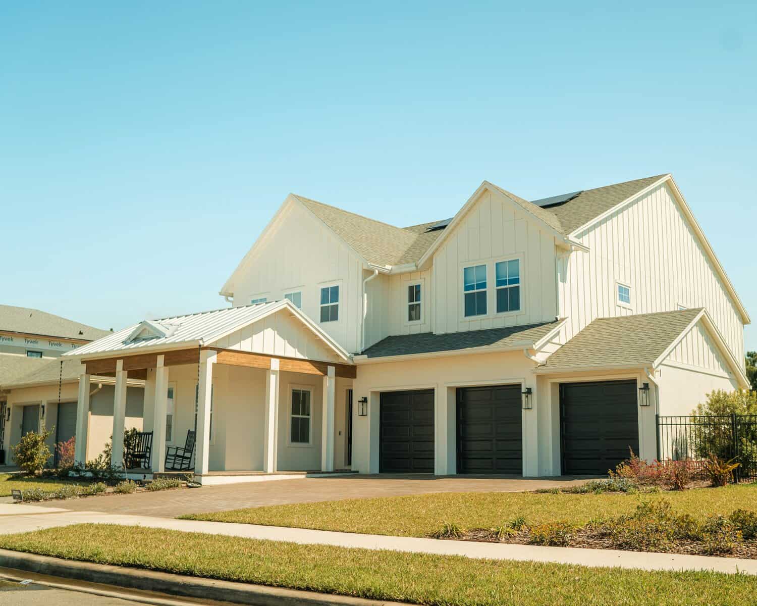 Two-story modern suburban house with a front porch, three black garage doors, white siding, and a well-kept lawn—designed to help protect against external power surges that can impact your home under a clear blue sky.