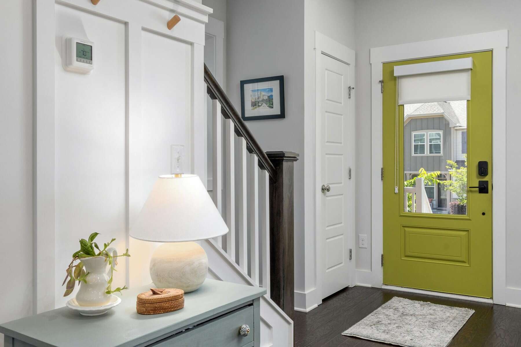 A modern entryway features a green front door, a staircase, a lamp on a cabinet protected from power surges, and a potted plant, with natural light streaming through the window.