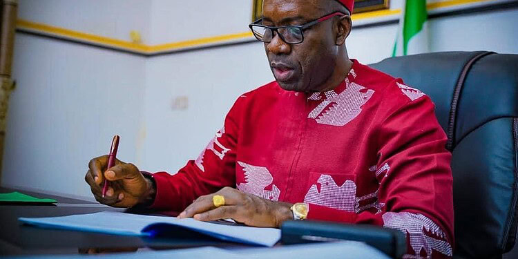 African man in traditional attire signing documents at a desk in an official setting with flags in the background.