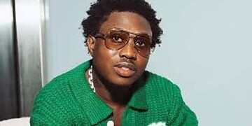 A young man with curly hair wearing sunglasses and a green sweater, looking confidently at the camera.