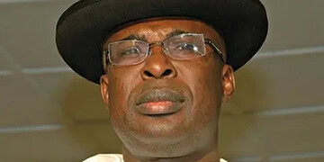 Photograph of a man wearing a black hat and glasses, looking serious in an indoor setting.