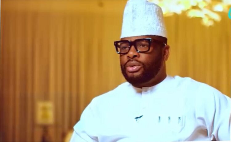 Professional Nigerian chef wearing traditional attire, glasses, and a hat, speaking confidently in a modern kitchen setting.