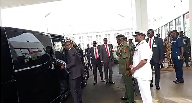 Black luxury vehicle being attended to by a man in a suit, surrounded by distinguished military and civilian officials at an official event, possibly a formal or diplomatic occasion.