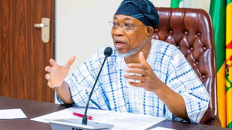 Nigeria government official speaking at meeting during press conference, wearing traditional attire and cap, seated at a desk with a microphone, behind national flags.