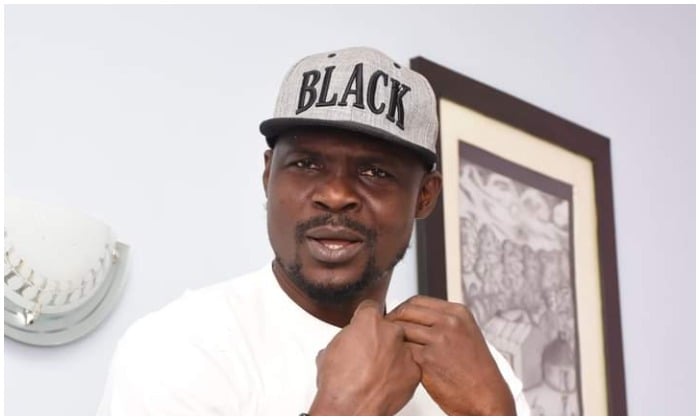 Black man wearing a grey cap with "BLACK" written on it, standing indoors with artwork on the wall behind him.