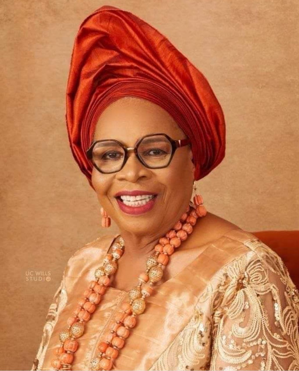 Vibrant Nigerian woman wearing traditional coral attire, gele headwrap, and glasses, smiling warmly. Embracing rich cultural fashion and elegance, showcasing ethnic pride and beauty. Ideal for features on African culture, fashion, and traditional accessories.