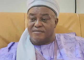 Elderly man wearing traditional attire and headwrap sitting on a beige sofa in a well-lit room, showcasing diverse cultural fashion.