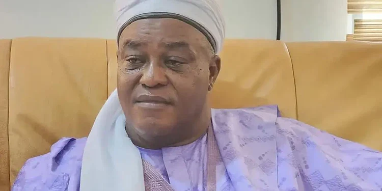 Elderly man wearing traditional attire and headwrap sitting on a beige sofa in a well-lit room, showcasing diverse cultural fashion.