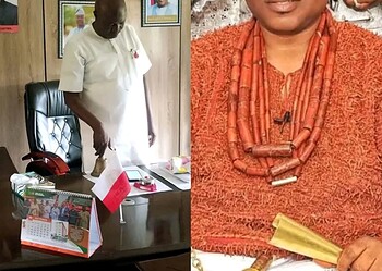 A government official in a white outfit standing in an office with framed photos and emblems on the wall, representing official political or administrative context in Nigeria.