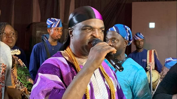 A leader or performer speaking into a microphone during a cultural event with traditional attire and music in the background.