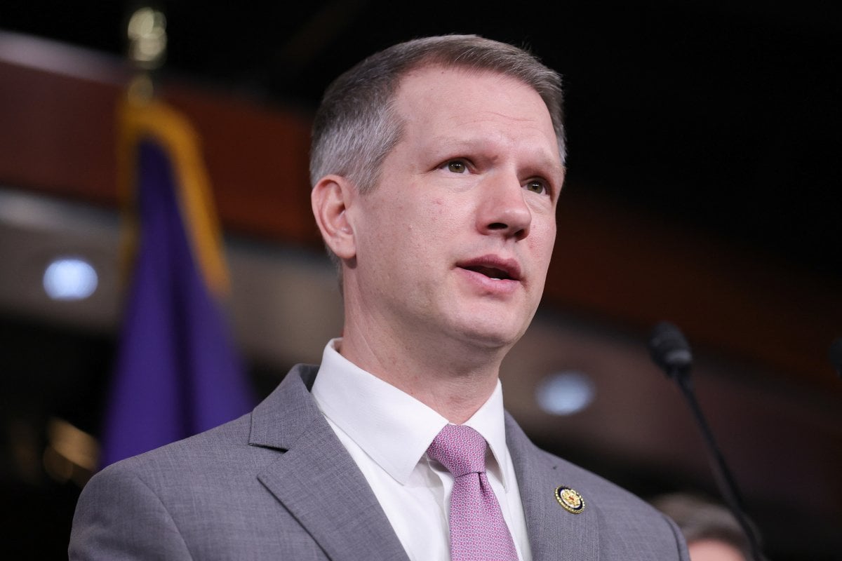 Esteemed politician speaking at a formal event, wearing a grey suit and a pink tie, with a purple flag in the background, highlighting politics, leadership, and government.