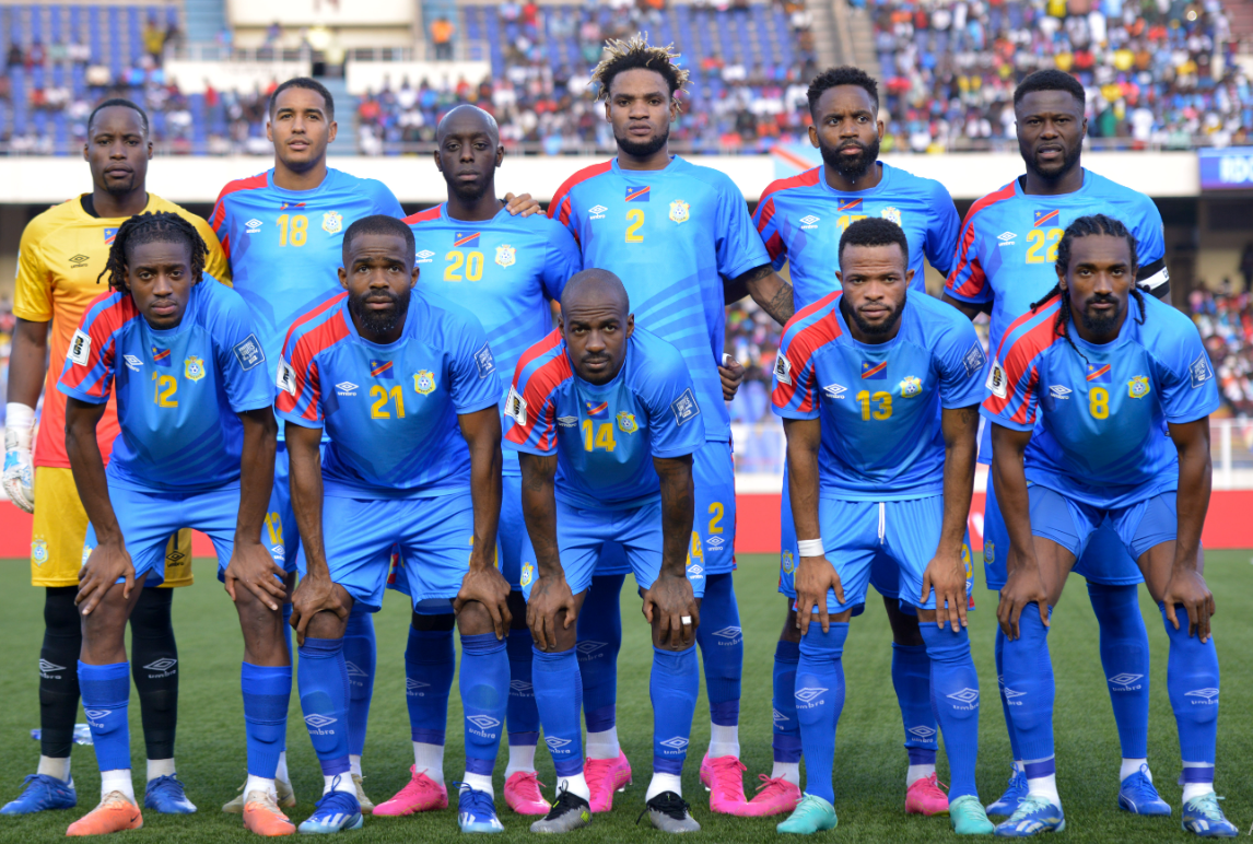 Vibrant team photo of soccer players in blue and yellow uniforms on the field before a match, showcasing team unity and diversity, with spectators in the background.