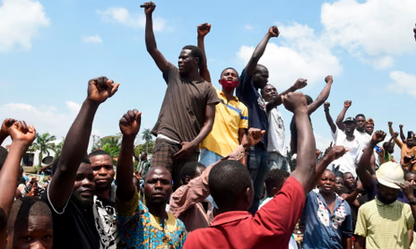 Protestors rallying outdoors, raising fists and voices for social justice, during a daytime demonstration with a cloudy sky background.