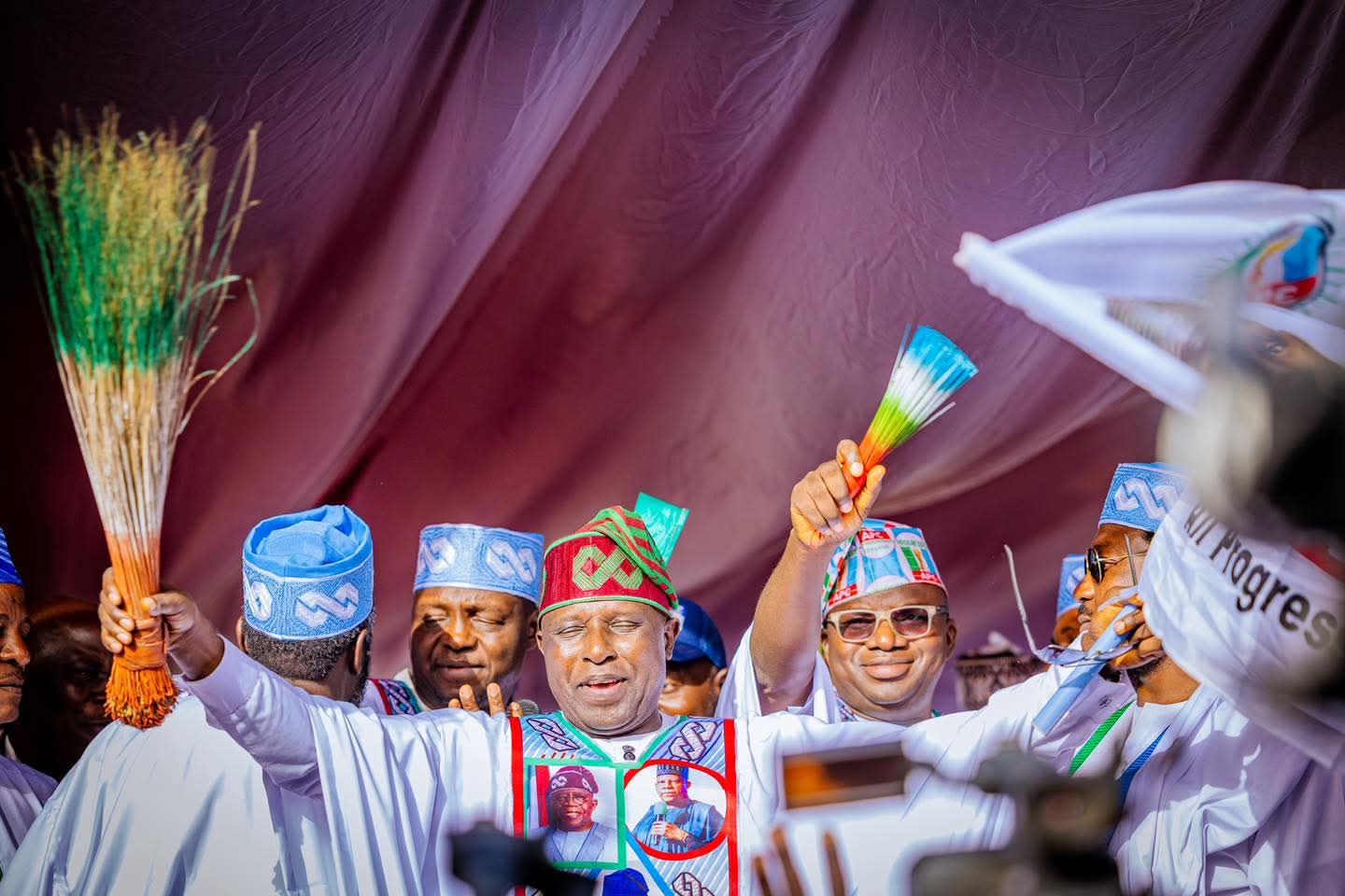 Colourful traditional Nigerian attire worn during a cultural celebration or festival, with men holding decorative fan-like objects under a vibrant canopy.