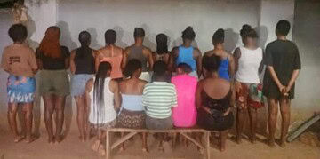 A group of people gathered outdoors at night, sitting and standing along a white wall, in a community setting.