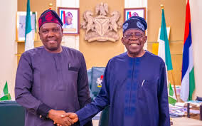A photo of two men in traditional African attire shaking hands in a formal setting with flags and a crest in the background.