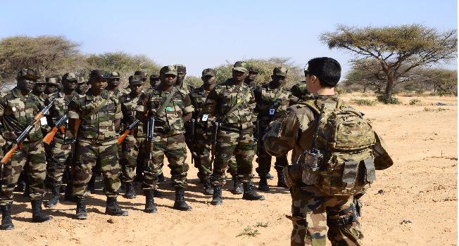 Soldiers in camouflage uniforms standing in formation listening to a commanding officer in a desert landscape.