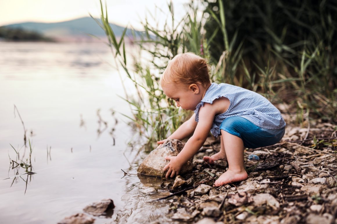 A small toddler girl playing outdoors by the river in summer.