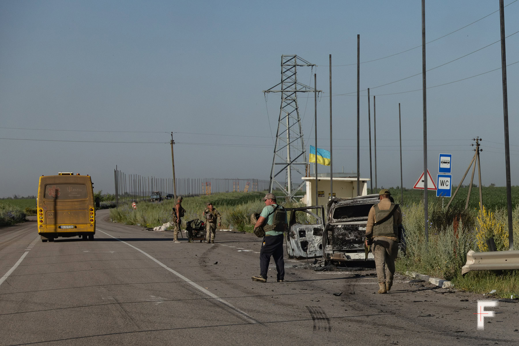 Спалене російським дроном авто й українські військові з мисливською зброєю, що охороняють робітників під час встановлення захисної сітки від дронів на трасі Краматорськ — Добропілля. Донецька область, 10 липня 2025 року. Андрій Дубчак / Frontliner.