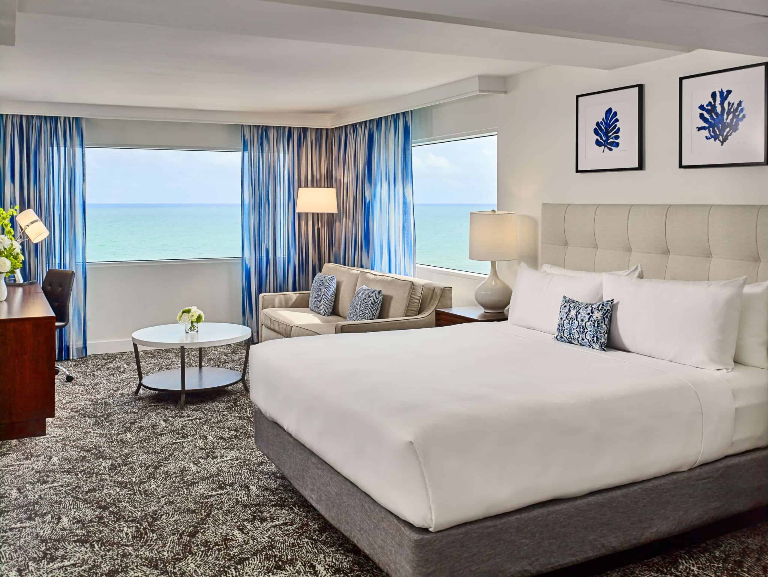 Hotel guestroom with a bed, seating area, working area, TV, and an ocean view.