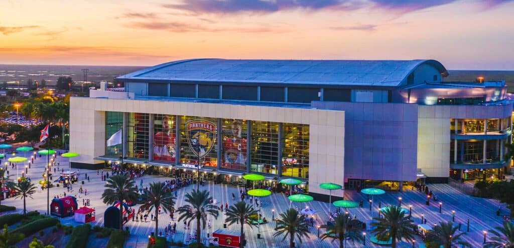 Exterior view of a large event arena at sunset with colorful lights and palm trees.