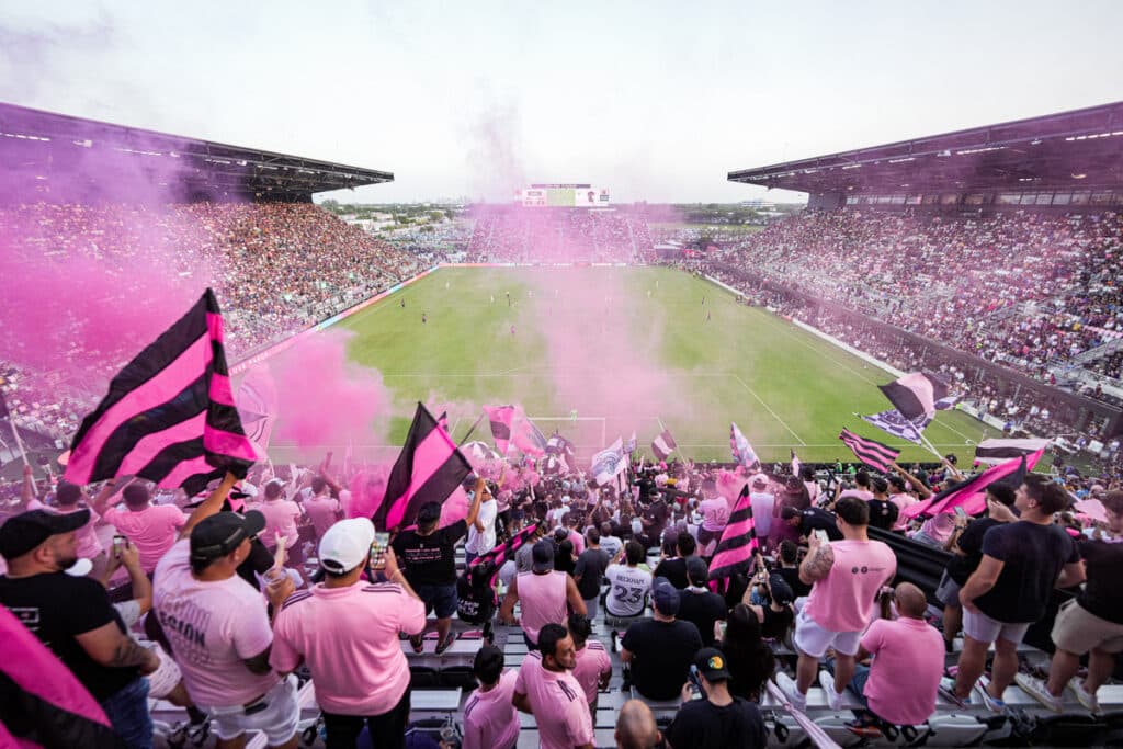 Excited crowd in a stadium with pink smoke and flags during a soccer match.