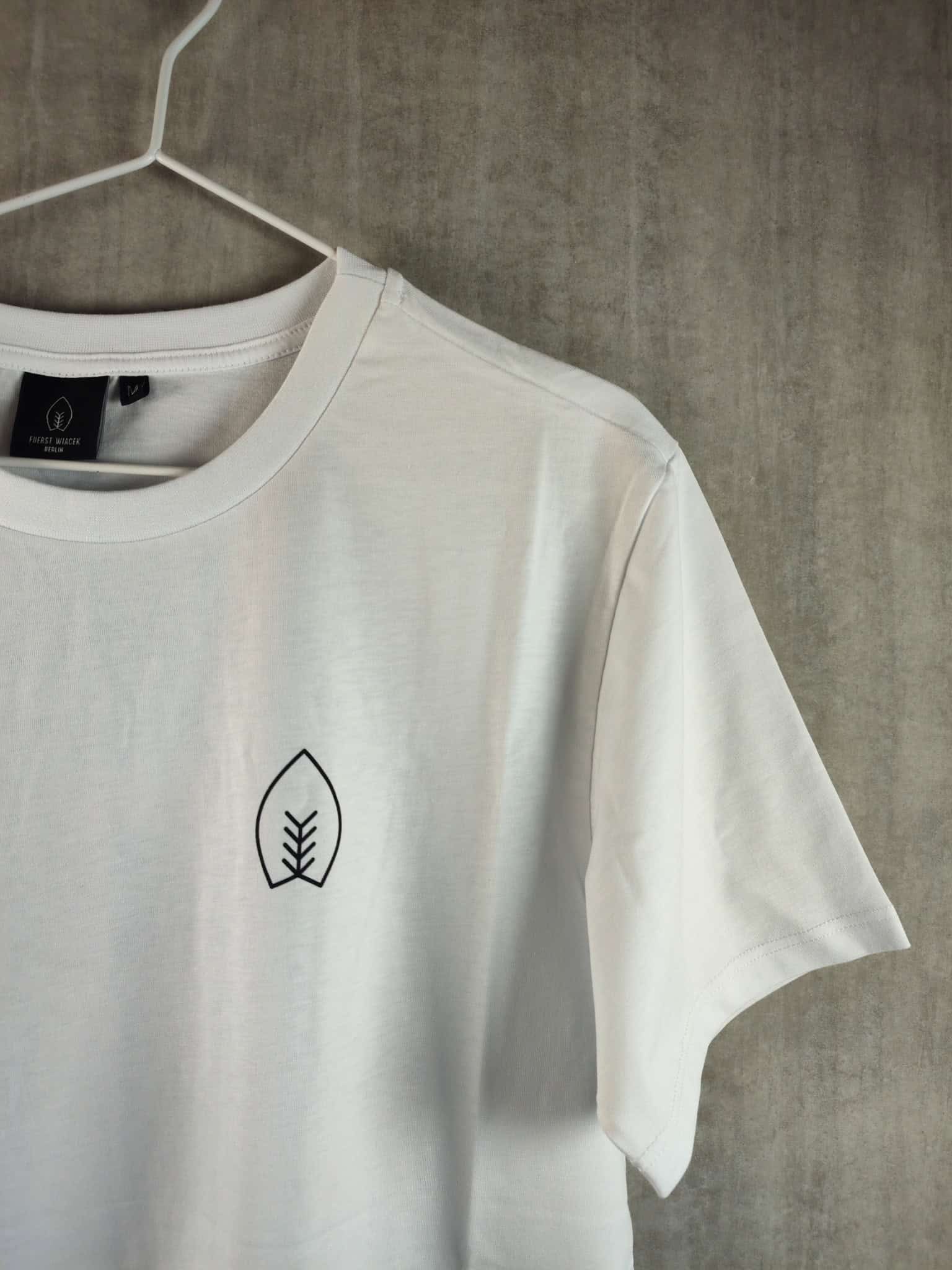 Minimalistisches weißes T-Shirt mit FUERST WIACEK-Logo, das auf einem weißen Bügel an einer Betonwand in der Brauerei hängt.