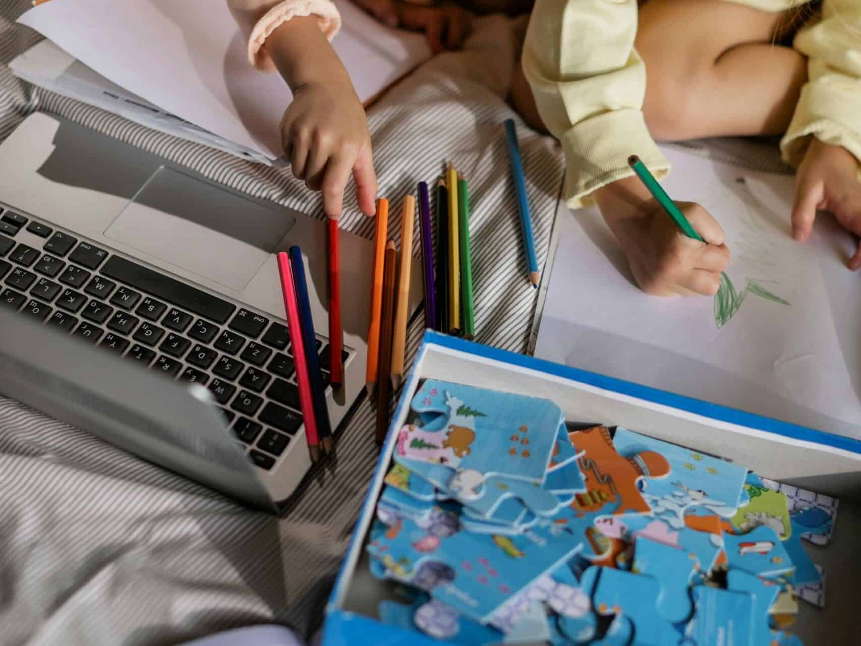 Two children drawing and solving puzzles at home, surrounded by colored pencils and a laptop.